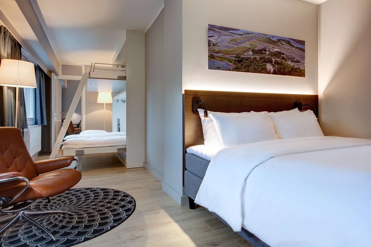 A hotel room with a bed, a chair, a desk, a lamp, and a painting on the wall.