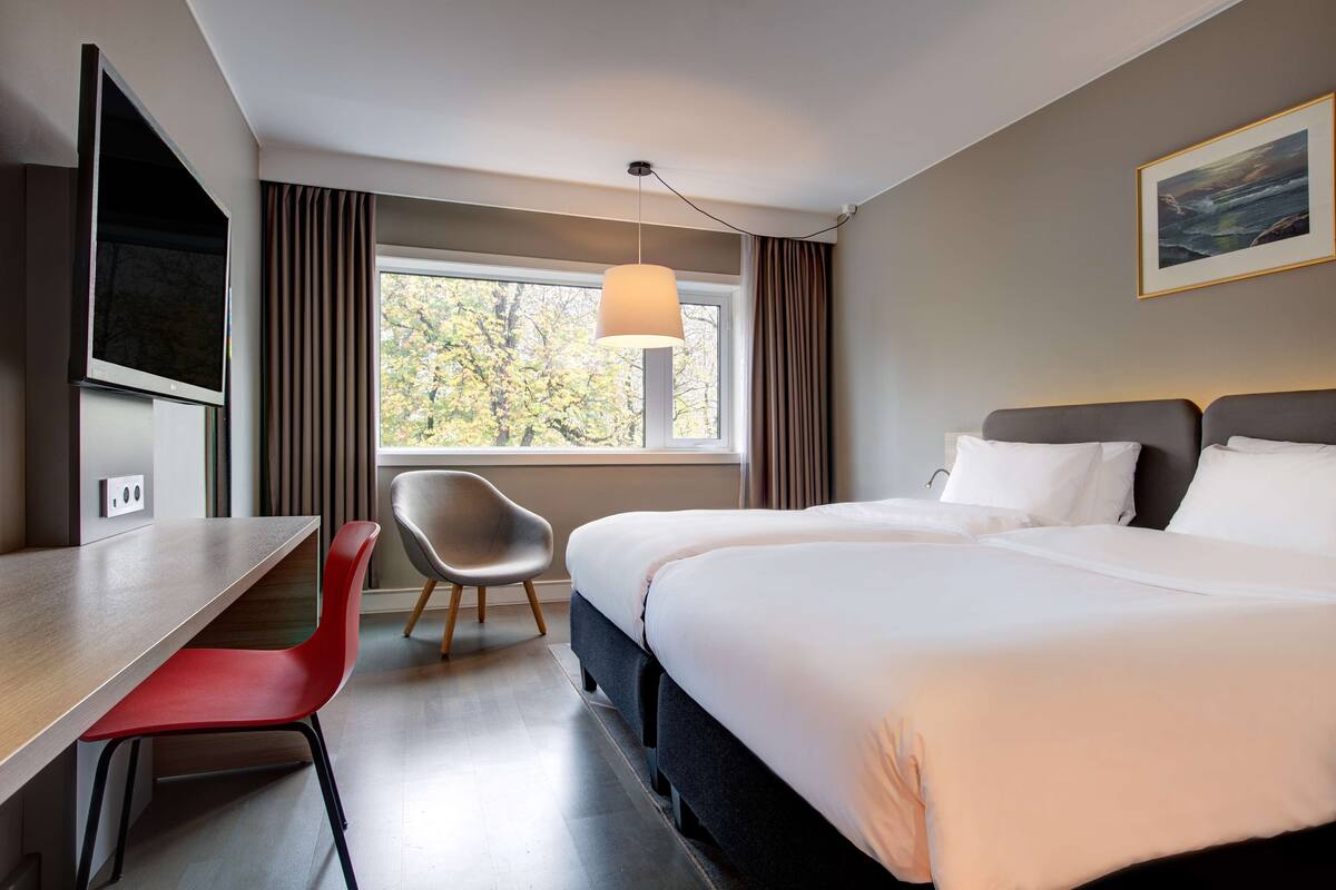A hotel room with a bed, a desk, a chair, a TV, and a window with curtains.