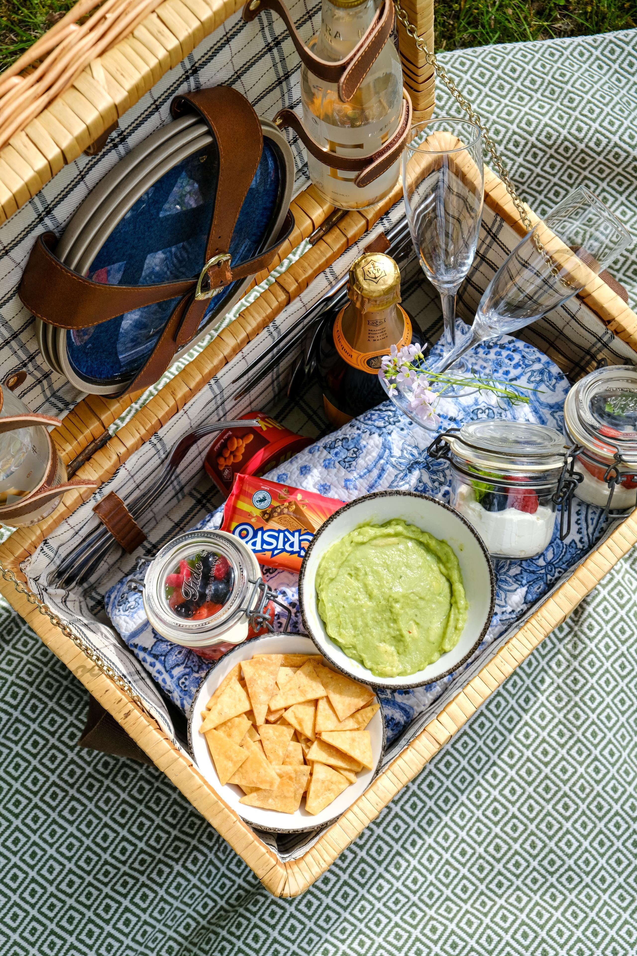 A picnic setup with a basket containing a bottle, wine glasses, a bowl of guacamole, chips, and a jar of salsa.
