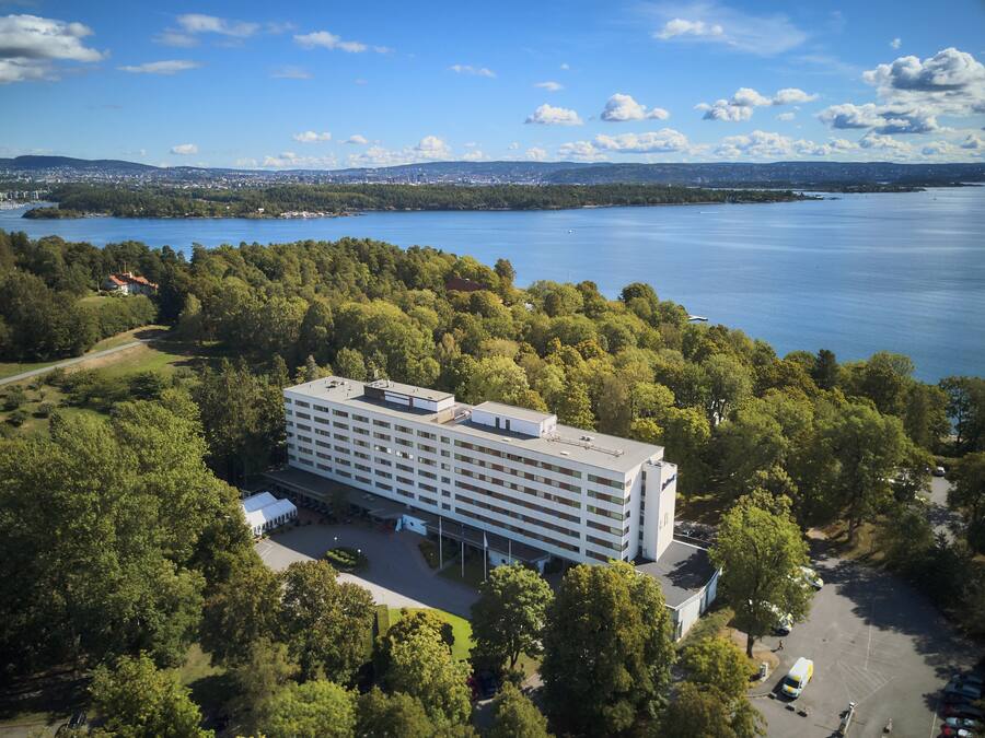 A modern hotel with a flat roof, surrounded by lush greenery and a lake.