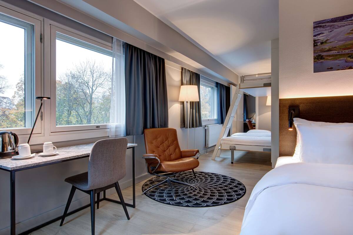 A modern hotel room with a desk, chair, bed, and a view of trees through large windows.