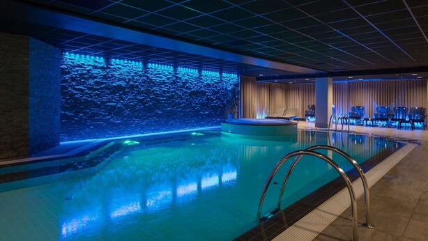 Una piscina interior moderna con una pared iluminada en azul y una zona de estar con vistas a la piscina.