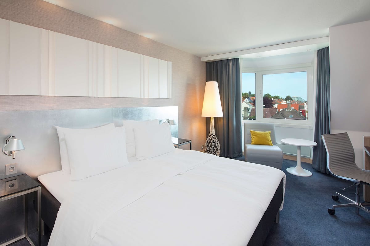 Ein Hotelzimmer mit einem großen Bett, einem Schreibtisch, einem Sessel und Blick auf die Stadt durch das Fenster.