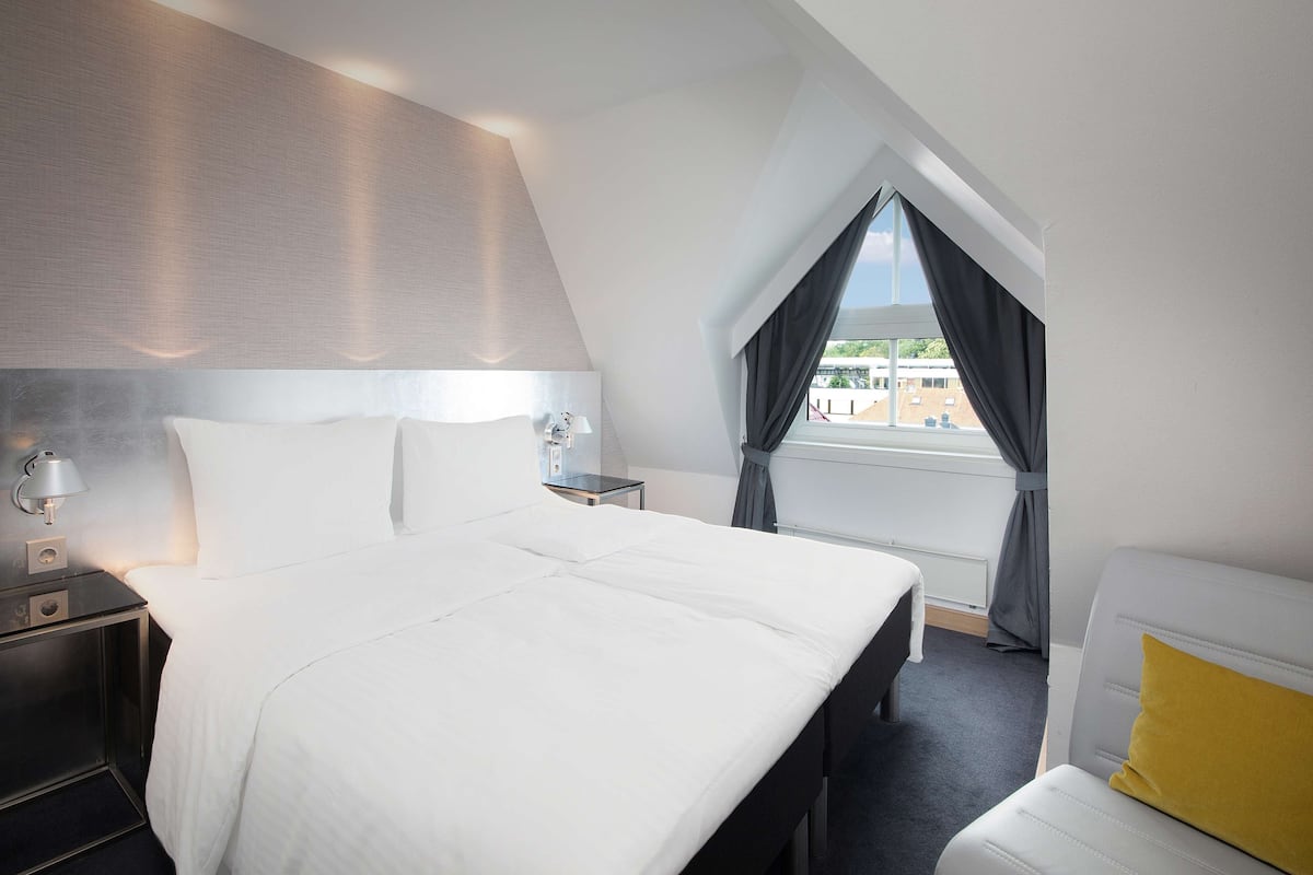 Ein modernes Hotelzimmer mit einem großen Bett, einem Nachttisch mit Lampe, einem kleinen Tisch und einem Fenster mit Vorhängen.