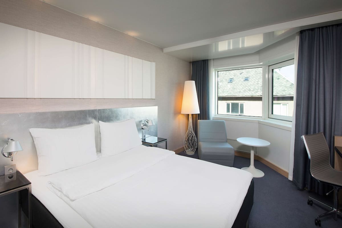 Ein Hotelzimmer mit einem großen Bett, einem Schreibtisch mit Lampe, einem Sessel, einem kleinen Tisch und einem Fenster mit Vorhängen.