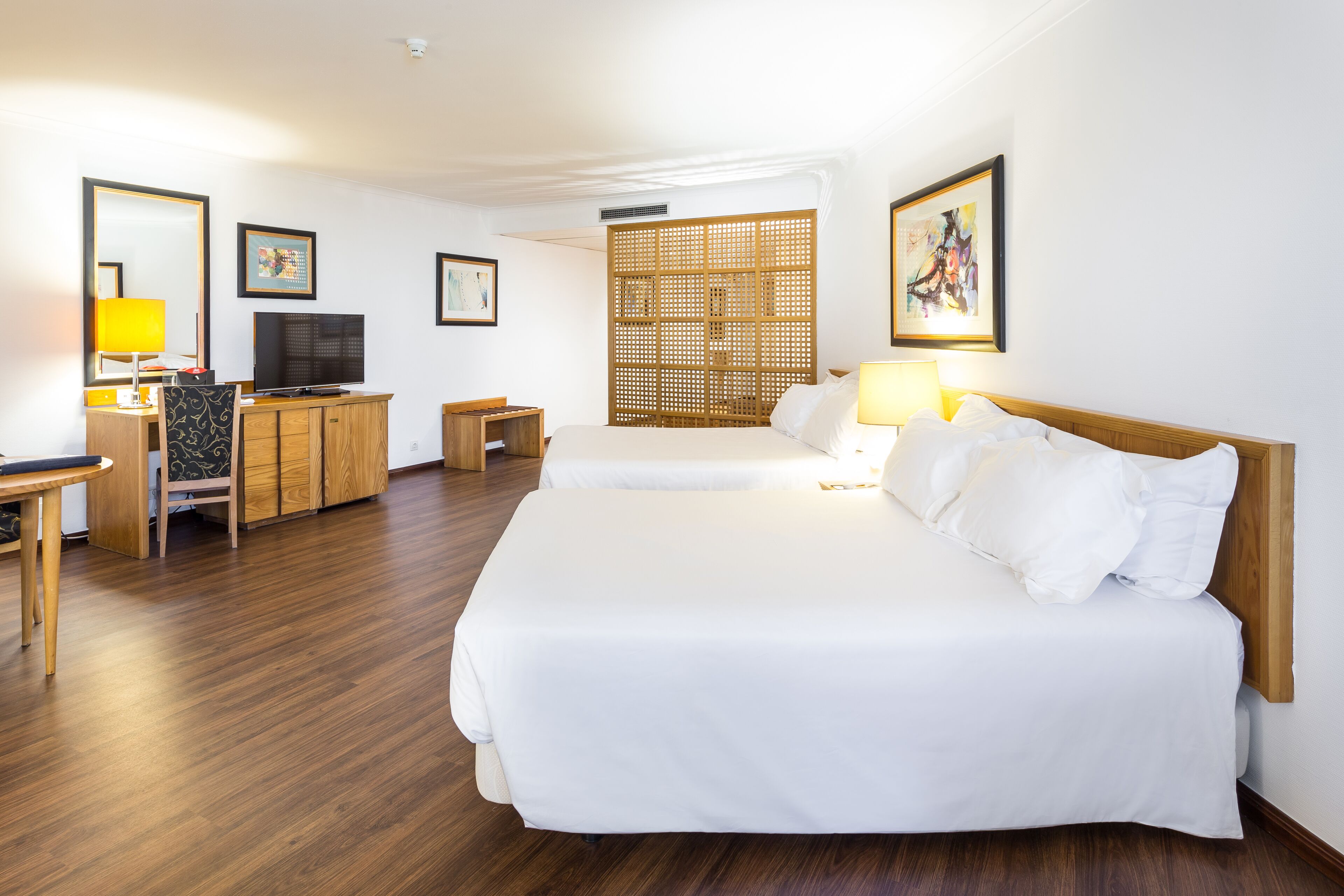 Habitación de hotel con dos camas, un escritorio con televisor, un armario de madera y un separador decorativo.