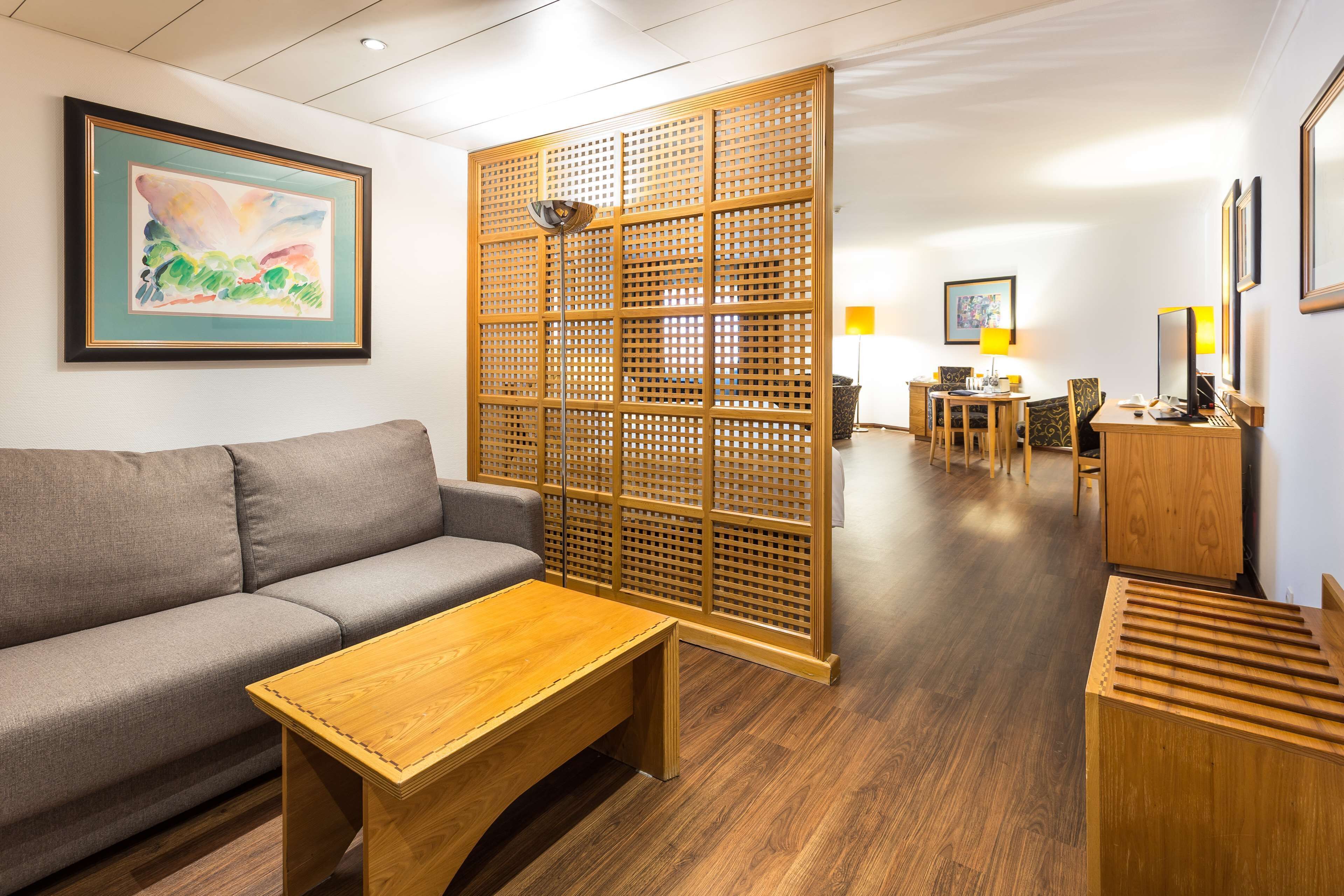 A modern hotel room with a wooden floor, a grey sofa, a wooden coffee table, a wooden partition, a desk with a chair, and a lamp.