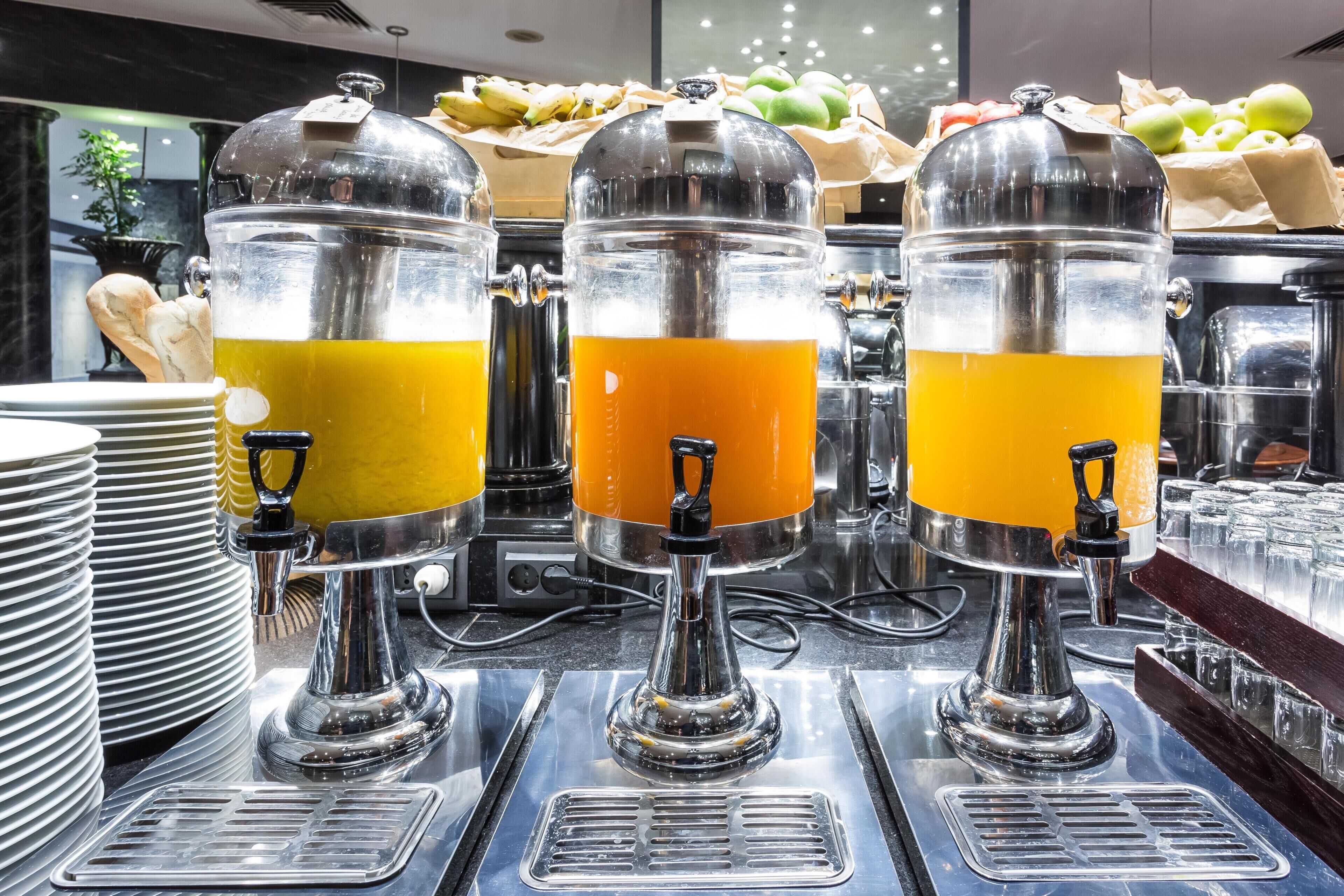 A buffet setup with three beverage dispensers, a fruit display, and stacked plates.