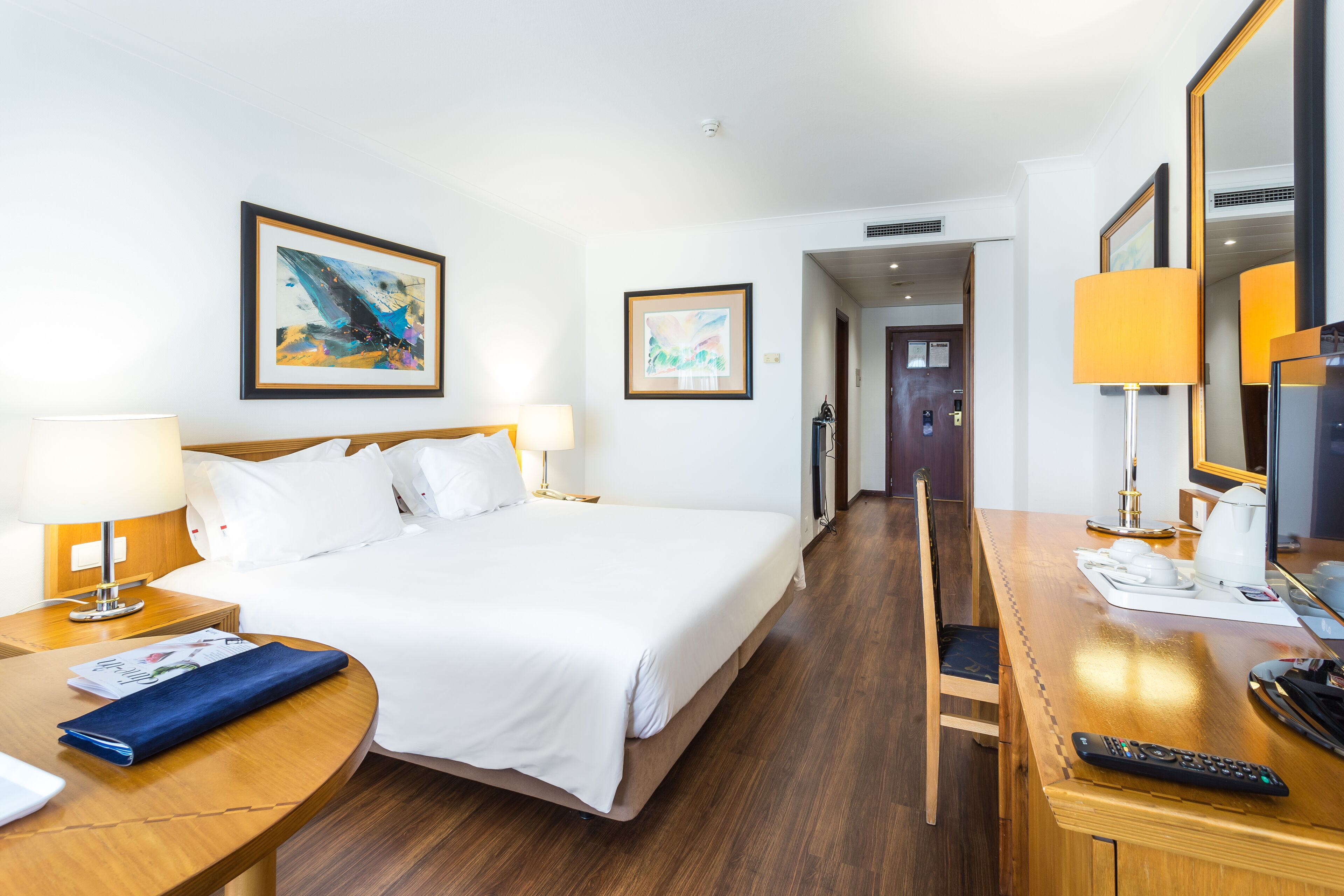 Habitación de hotel con una cama grande, un escritorio con lámpara, una silla y un televisor. Se observan cuadros enmarcados en la pared y una puerta que da a otra habitación.