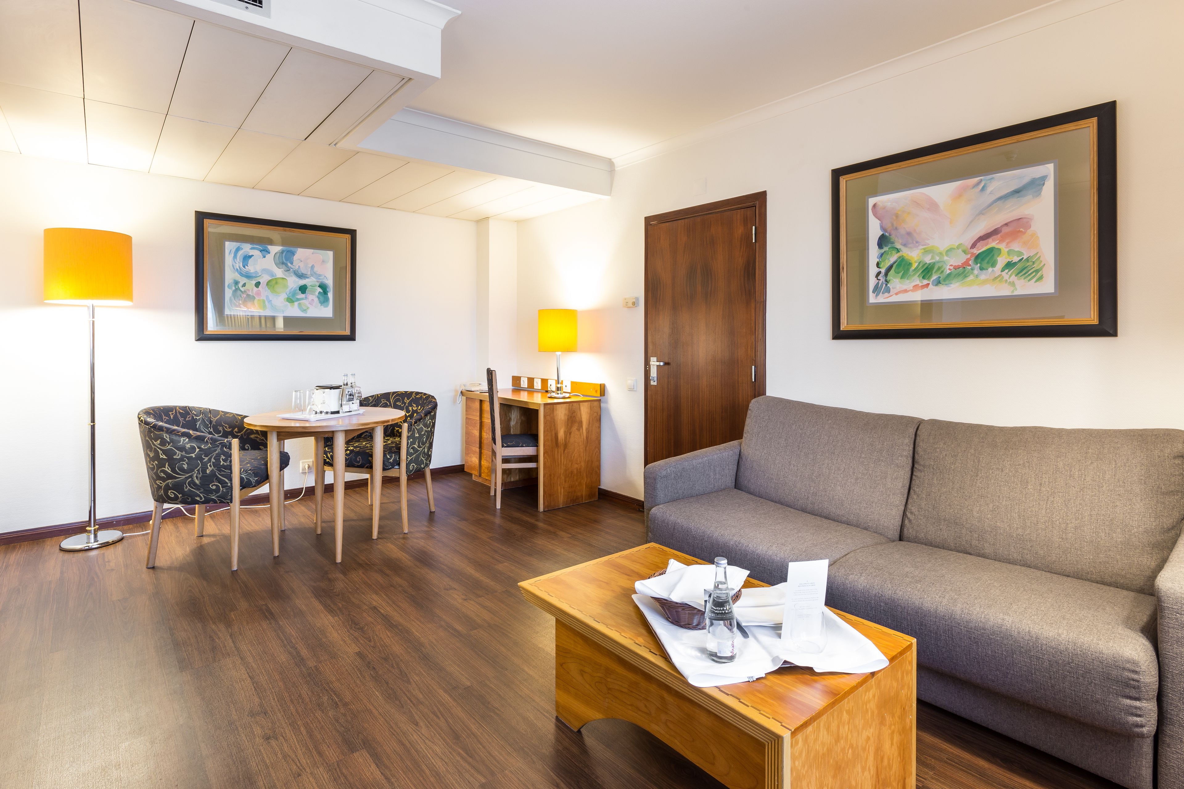 A hotel room with a sofa, a dining table with chairs, a lamp, and two framed artworks on the wall.