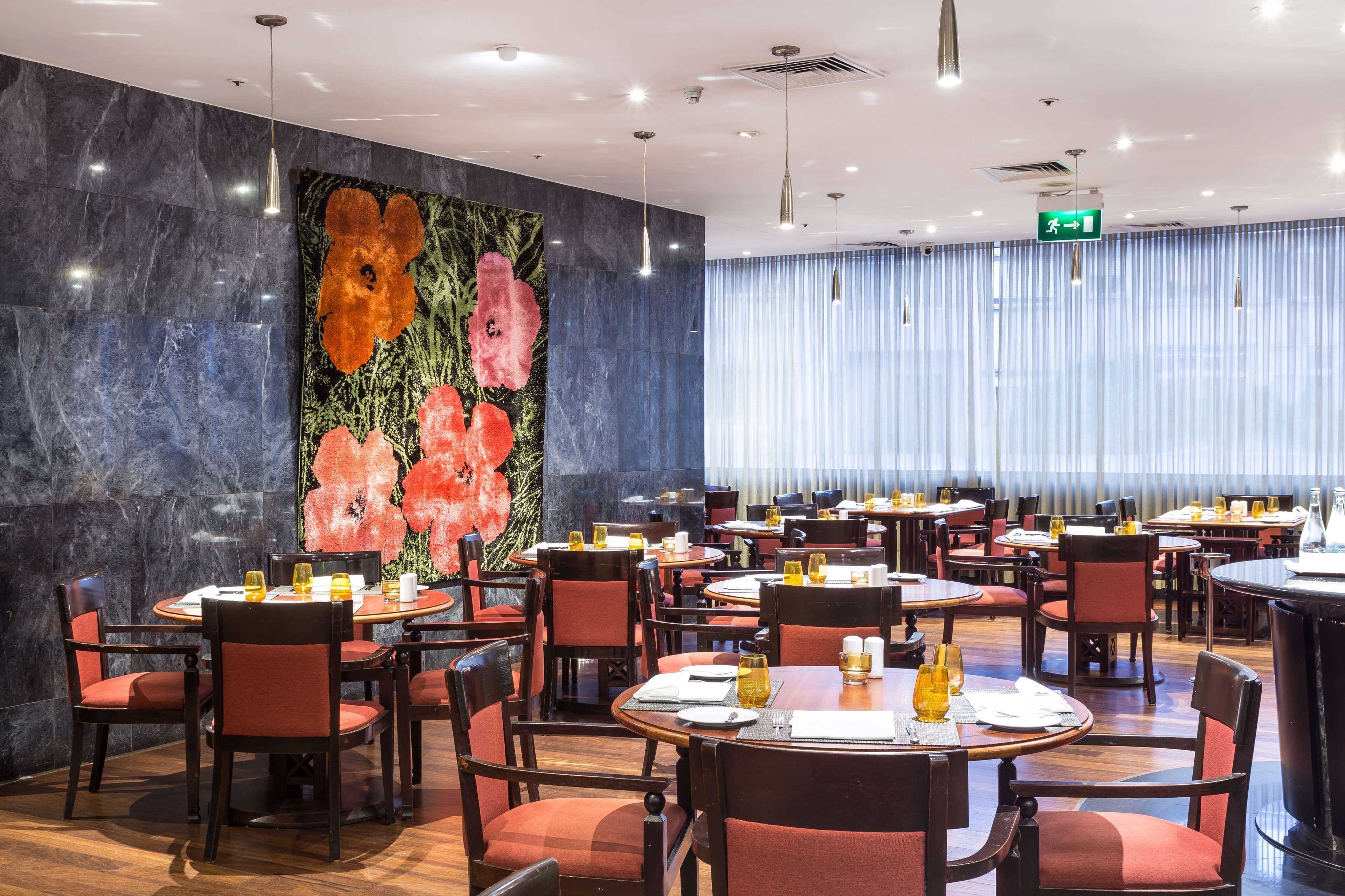 Un restaurante moderno con mesas redondas y sillas rojas, un gran mural floral en la pared y vistas de la ciudad a través de amplias ventanas.