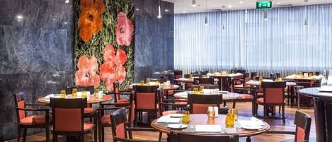 A modern restaurant with round tables and red chairs, a large floral mural on the wall, and a view of the city through large windows.