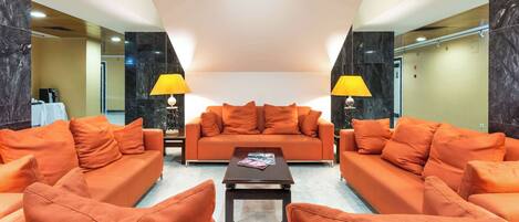A modern hotel lobby with a central orange sofa, two side sofas, a coffee table, and two lamps.