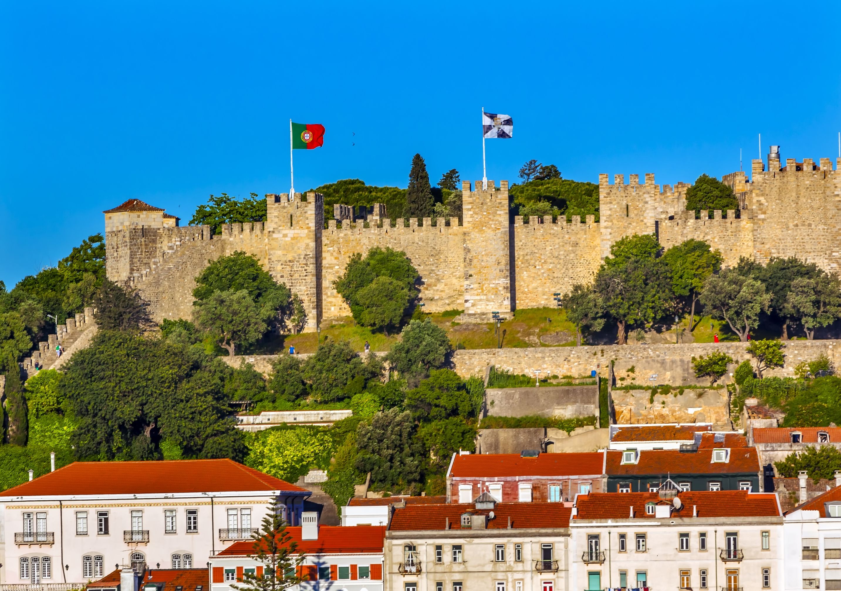 A historic stone castle with flags, surrounded by lush greenery and residential buildings with red-tiled roofs.