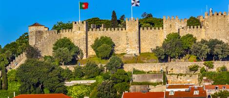 Un château de pierre historique, orné de drapeaux, entouré d’une végétation luxuriante et bordé de bâtiments résidentiels aux toits de tuiles rouges.