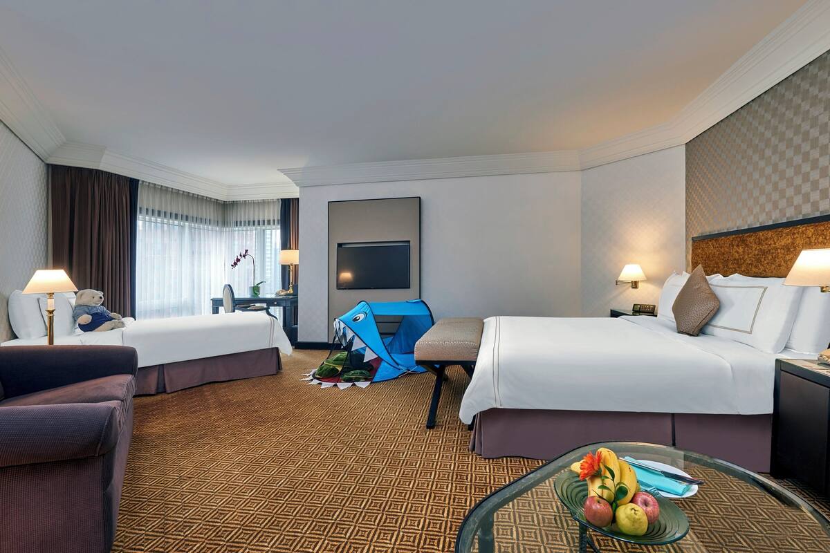 A hotel room with two beds, a sofa, a TV, and a glass table with fruit.
