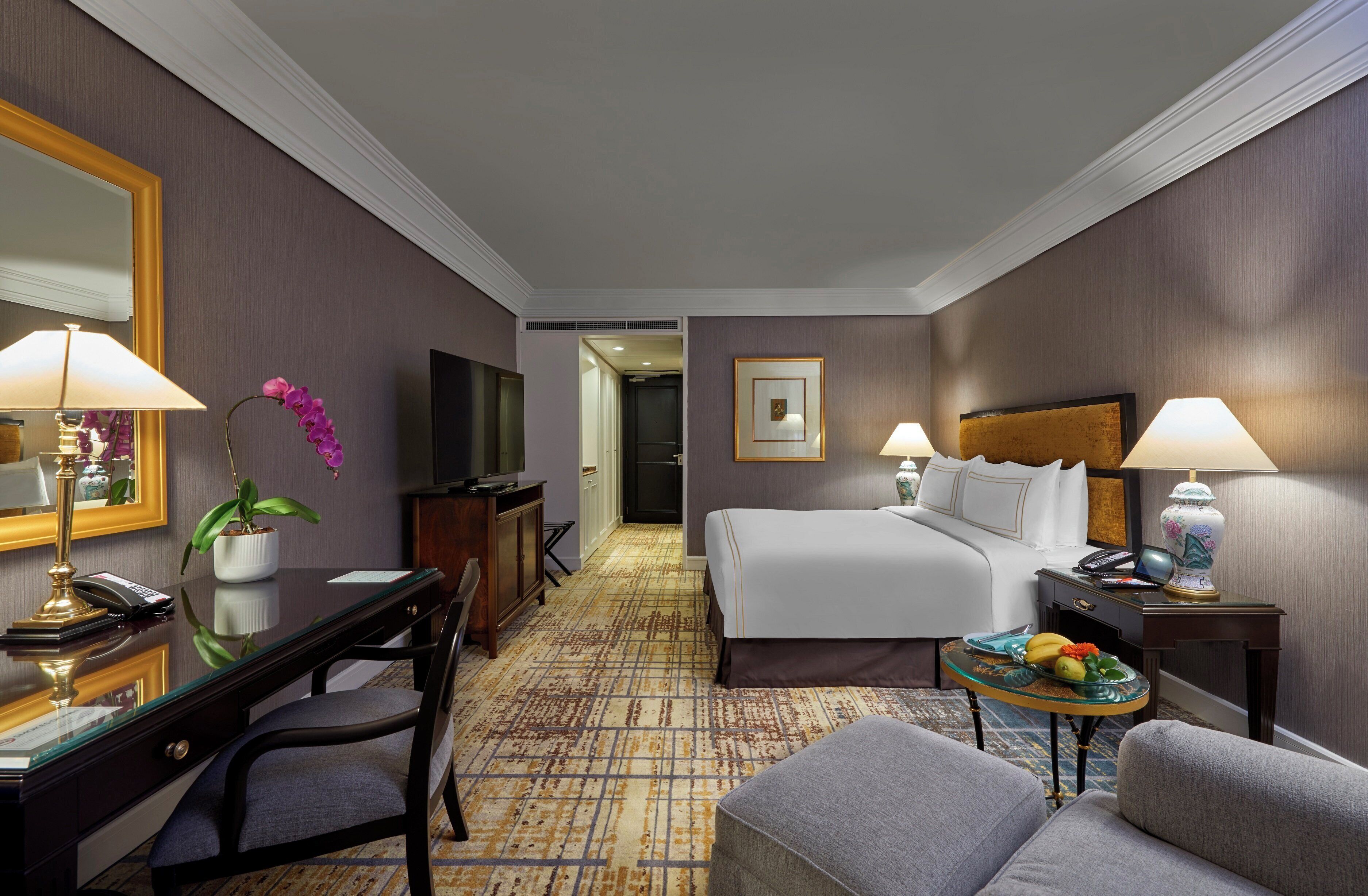 A hotel room with a bed, desk, chair, and a small table with fruit.