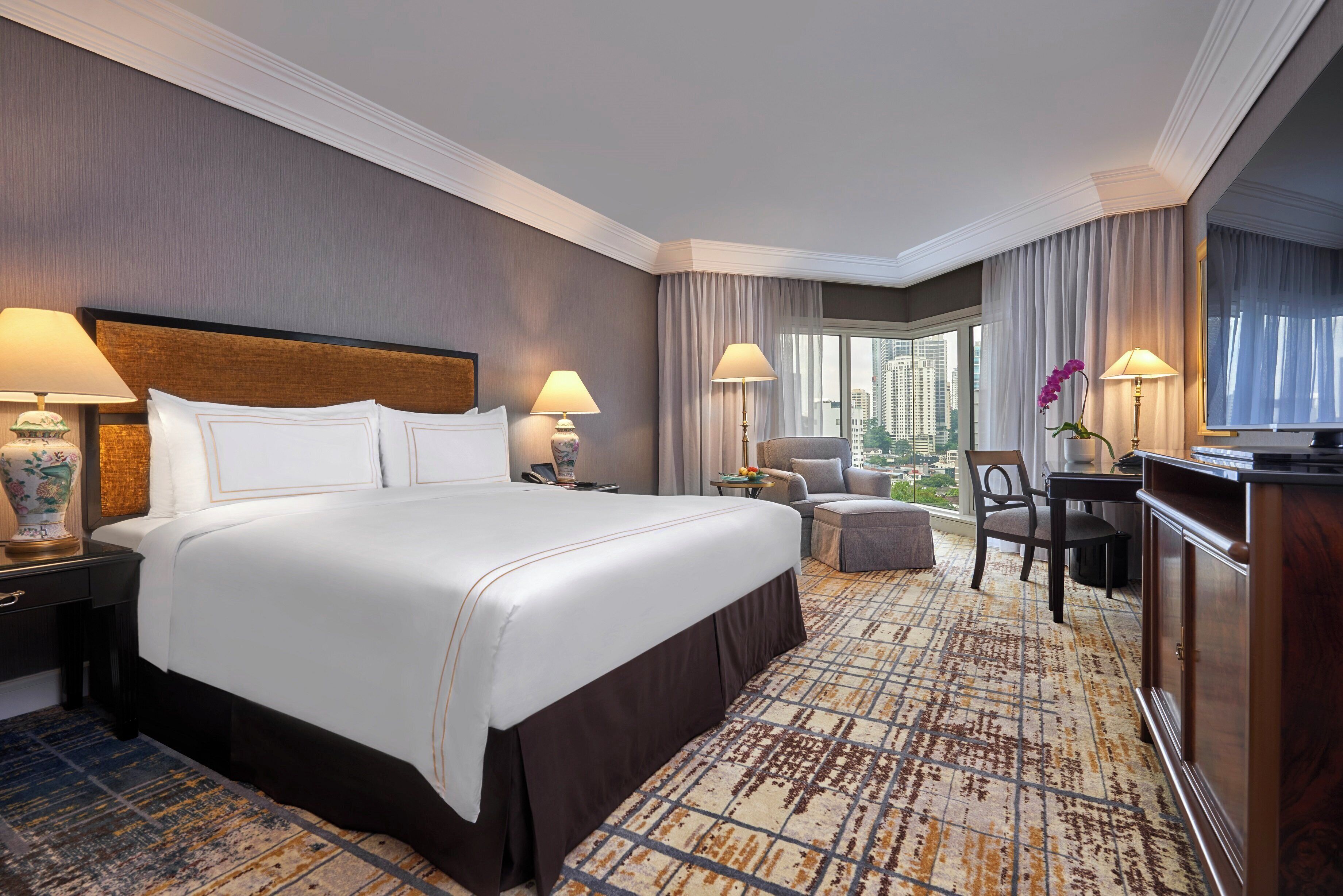 A hotel room with a large bed, two bedside tables with lamps, a chair, a desk, and a view of the city.