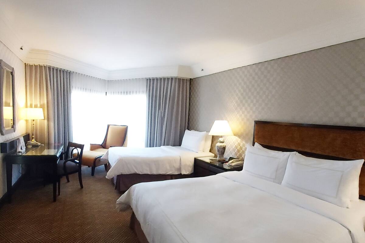 A hotel room with two beds, a desk, a chair, and a lamp.