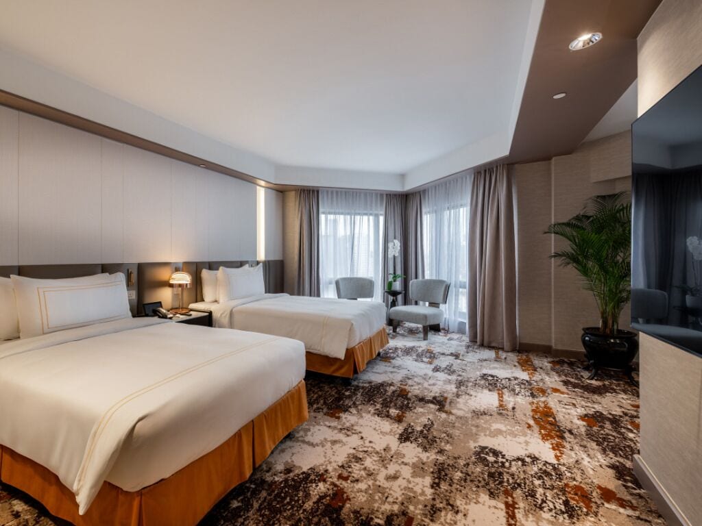 A hotel room with two beds, a desk, a chair, a potted plant, and a large window with curtains.