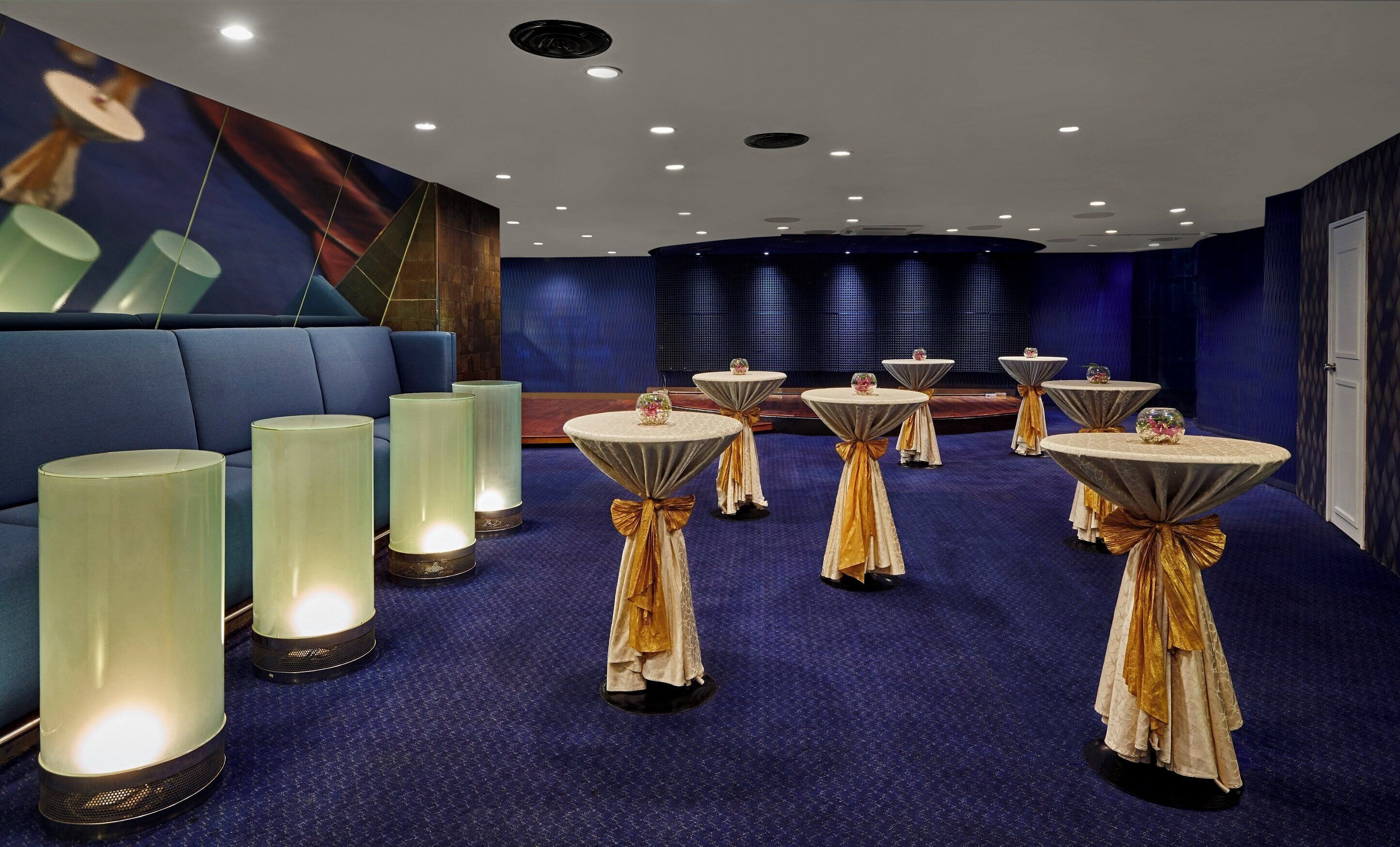 Un hall d’hôtel avec des tables rondes, de grandes lampes sur pied et une moquette bleue.