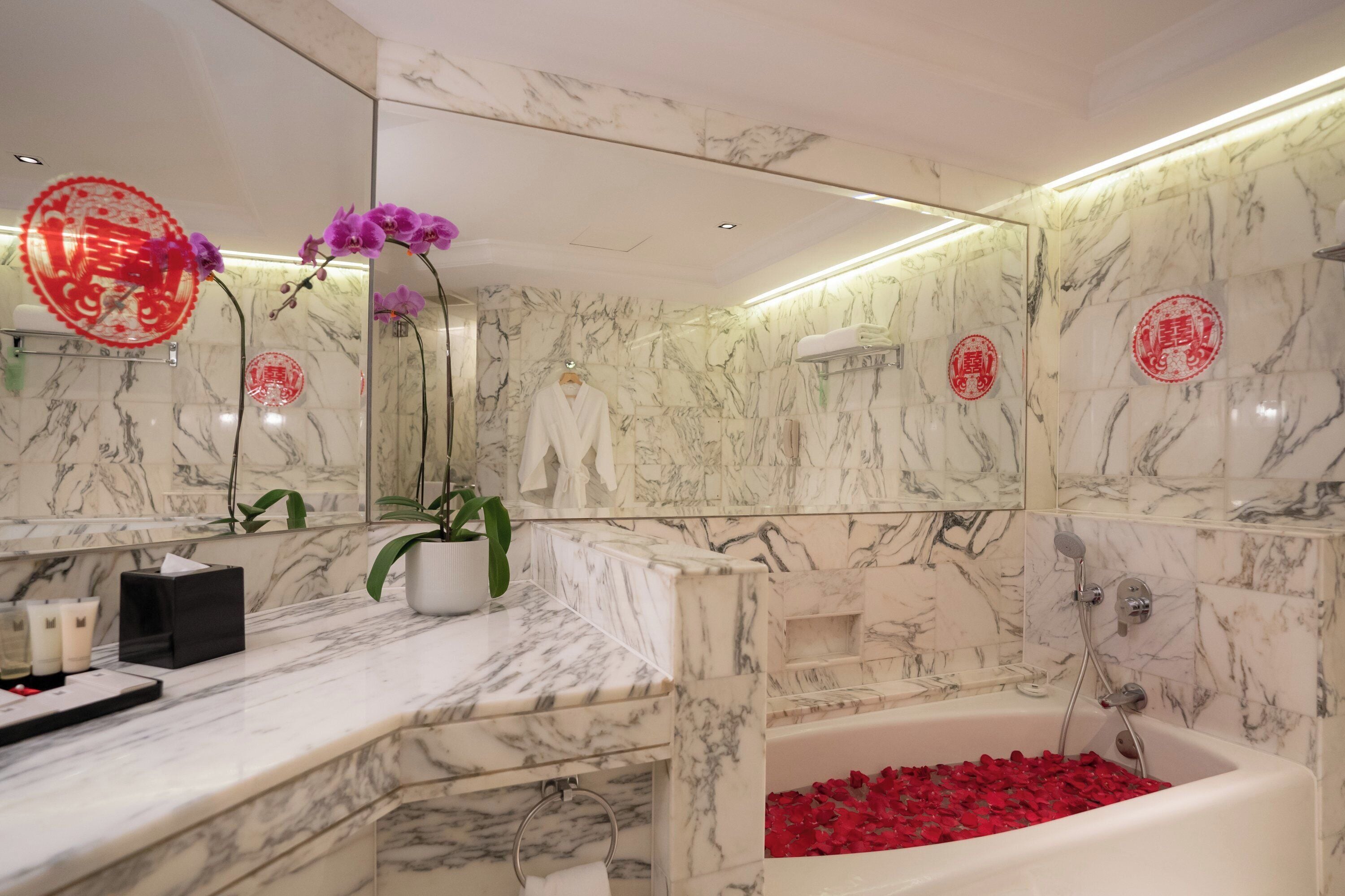 A bathroom with marble walls, a bathtub filled with rose petals, a towel rack, and a potted plant.