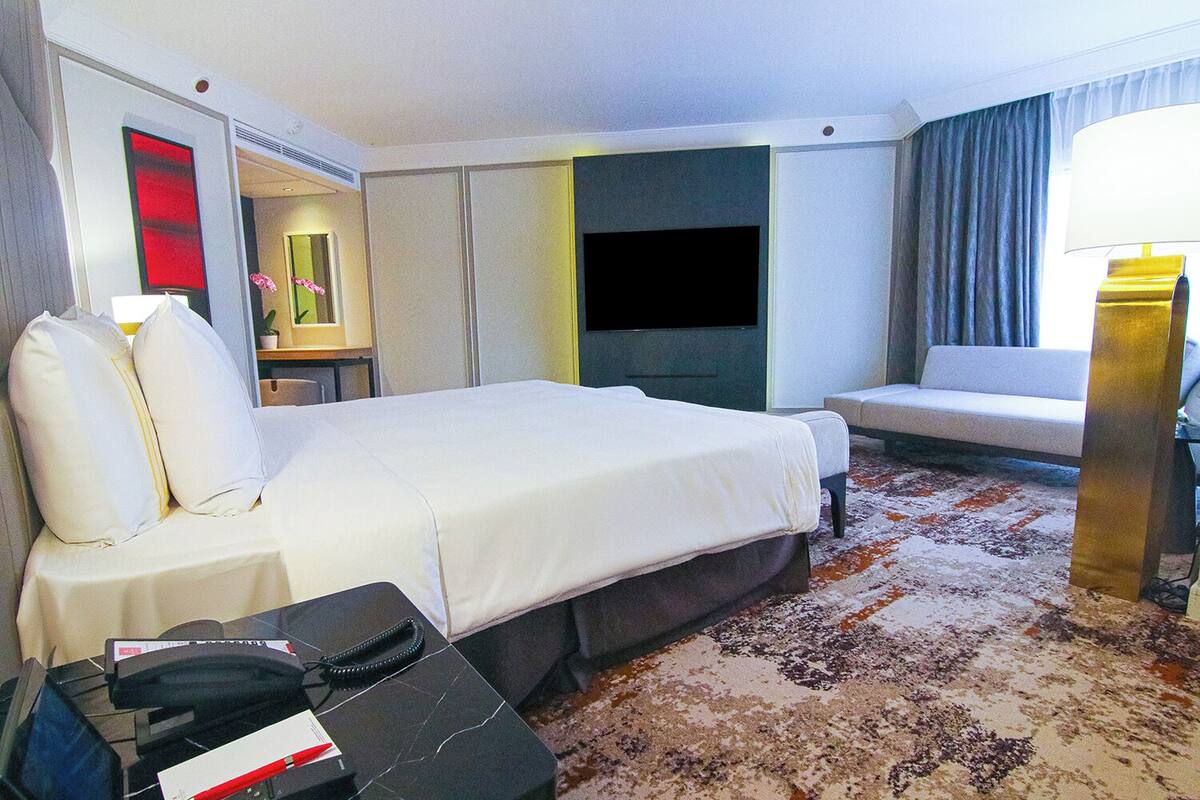A hotel room with a bed, a desk with a telephone, a TV, a sofa, and a lamp.