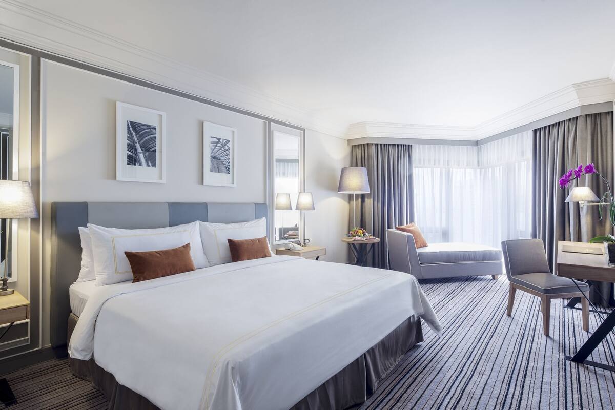 A hotel room with a large bed, a desk with a chair, a small table with a lamp, and a large window with curtains.