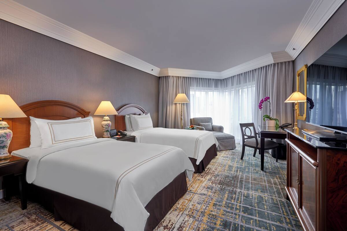 A hotel room with two beds, a desk, a chair, a TV, and a large window with curtains.