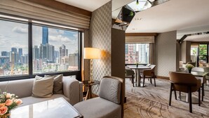 A hotel room with a city view, a sofa, a dining table, and a lamp.