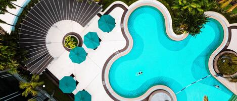An aerial view of a swimming pool with a unique wavy design, surrounded by palm trees and shaded by umbrellas.