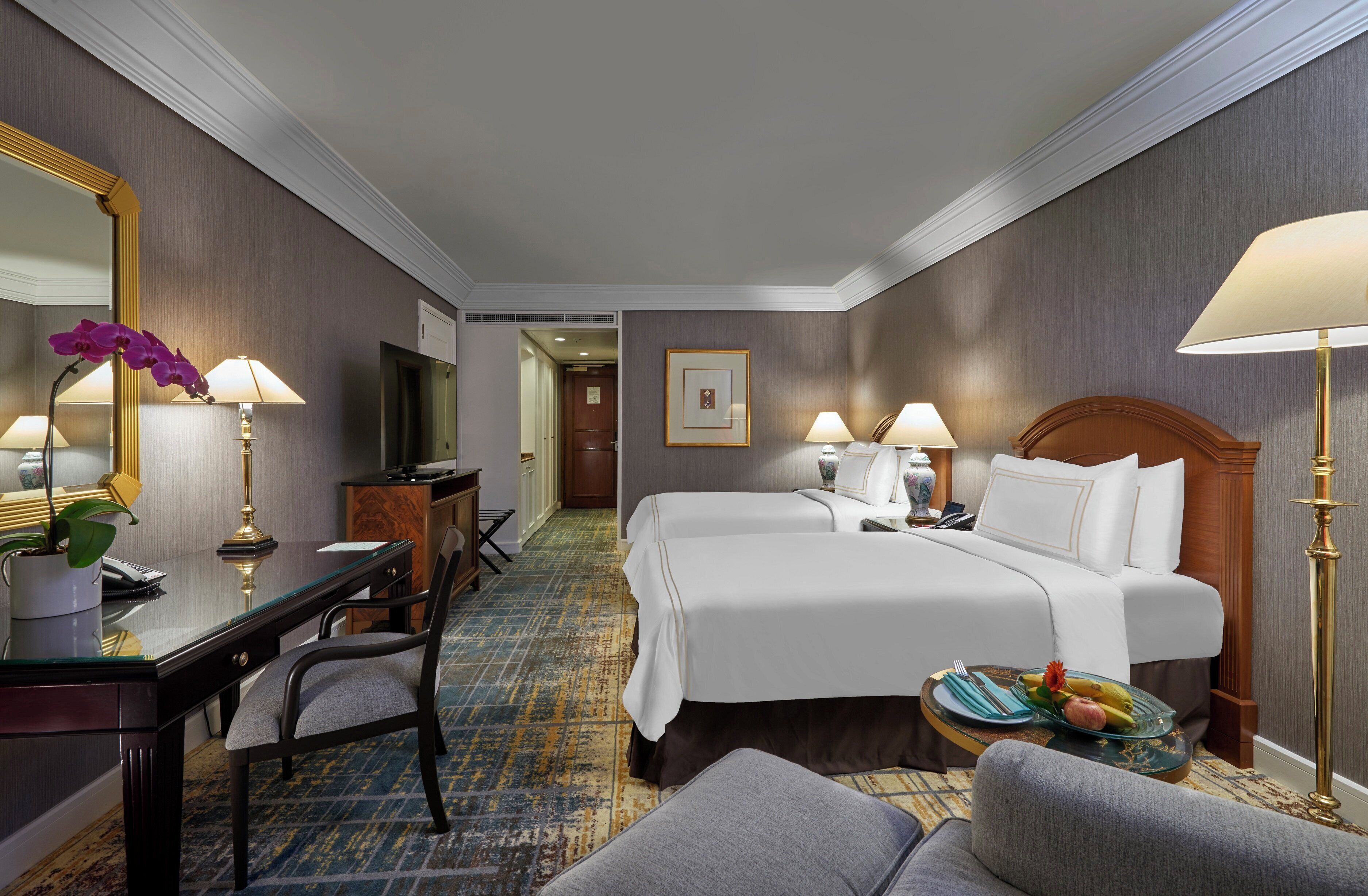 Habitaci ón de hotel con dos camas, un escritorio, una silla, televisión y una mesa de comedor con una bandeja de frutas.