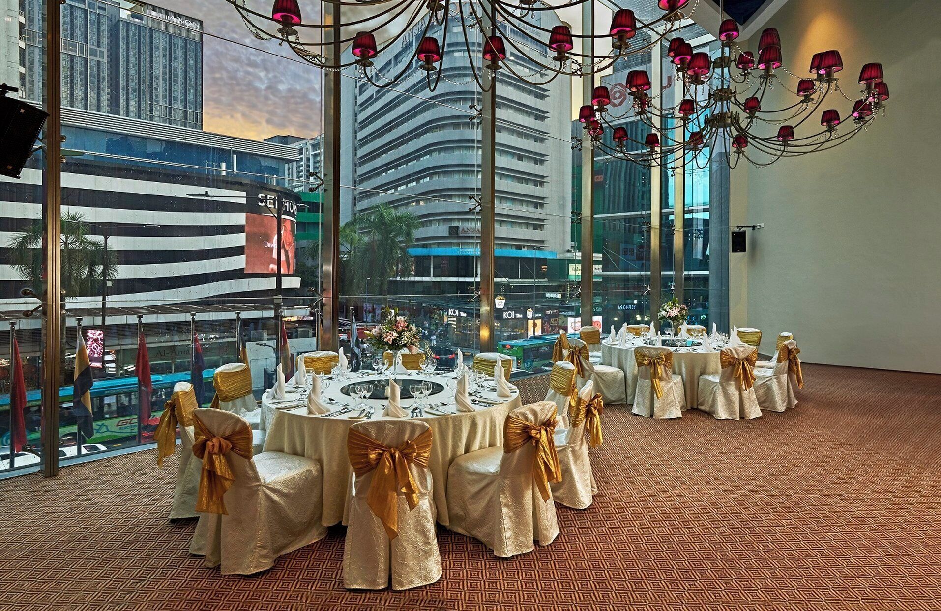 A banquet hall with round tables set for events, featuring gold sashes on chairs and a chandelier with red lampshades.
