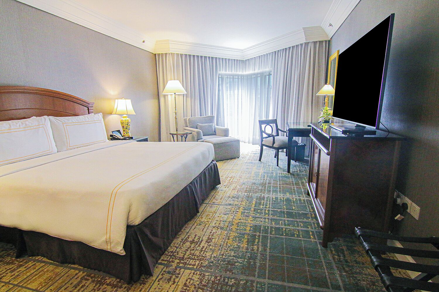 A hotel room with a large bed, a desk with a flat-screen TV, two chairs, a lamp, and a large window with curtains.