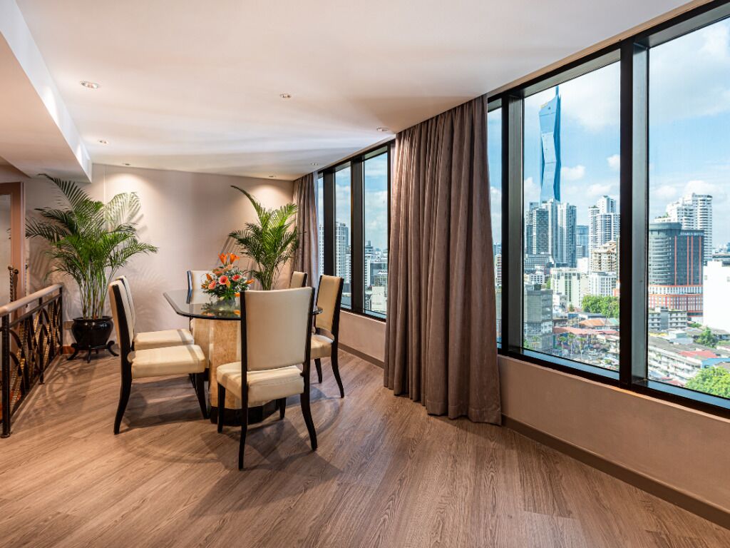 A modern hotel room with a large window, a dining area with a table and chairs, and potted plants.