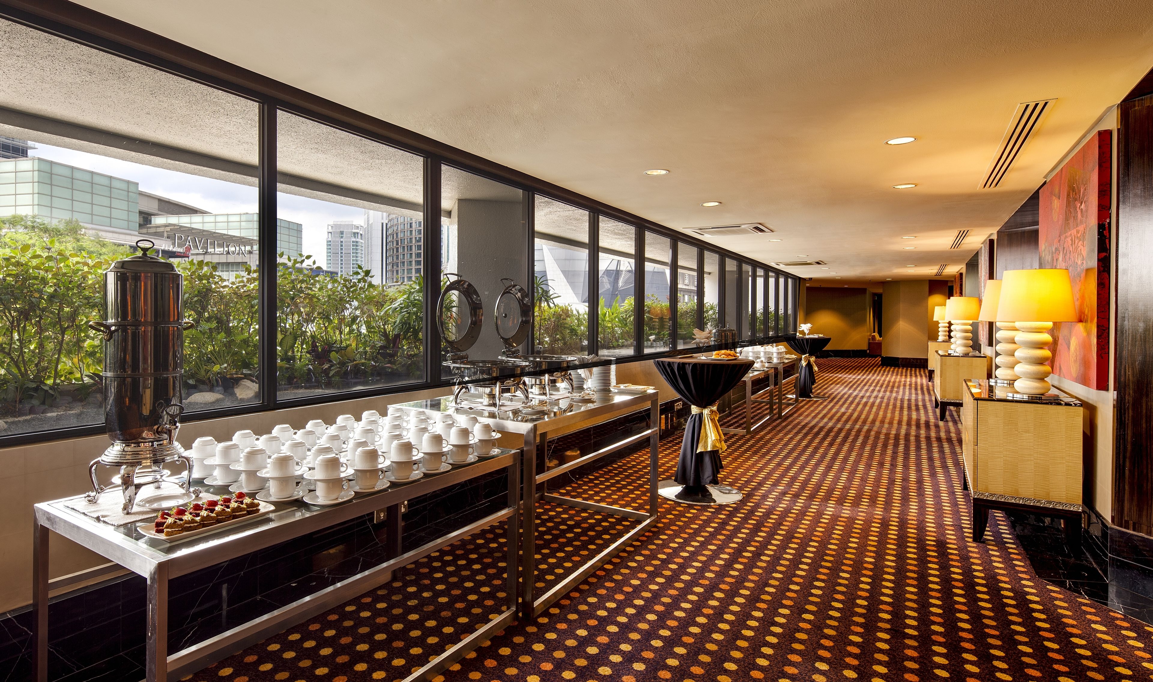 A hotel room with a buffet setup, large windows with a city view, and a patterned carpet.