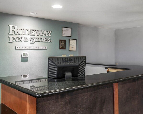 Un comptoir d’accueil doté d’un plan de travail foncé et d’un revêtement mural en bois. Au-dessus du comptoir, on peut lire une enseigne indiquant : « RODEWAY INN & SUNTES BY CHOICE HOTELS ».