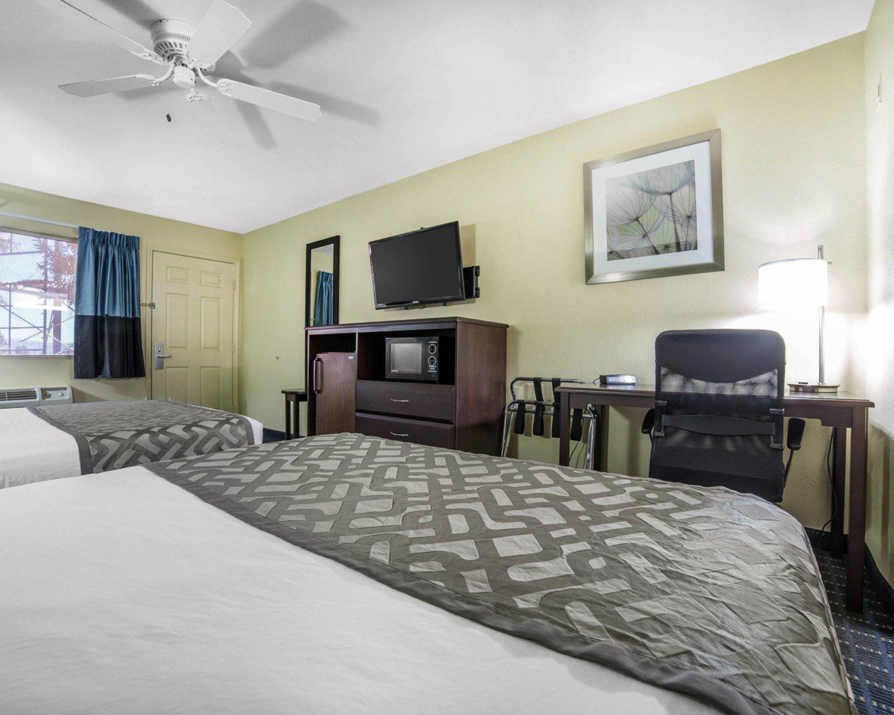 A hotel room with two beds, a television, a desk with a chair, a ceiling fan, and a window with curtains.