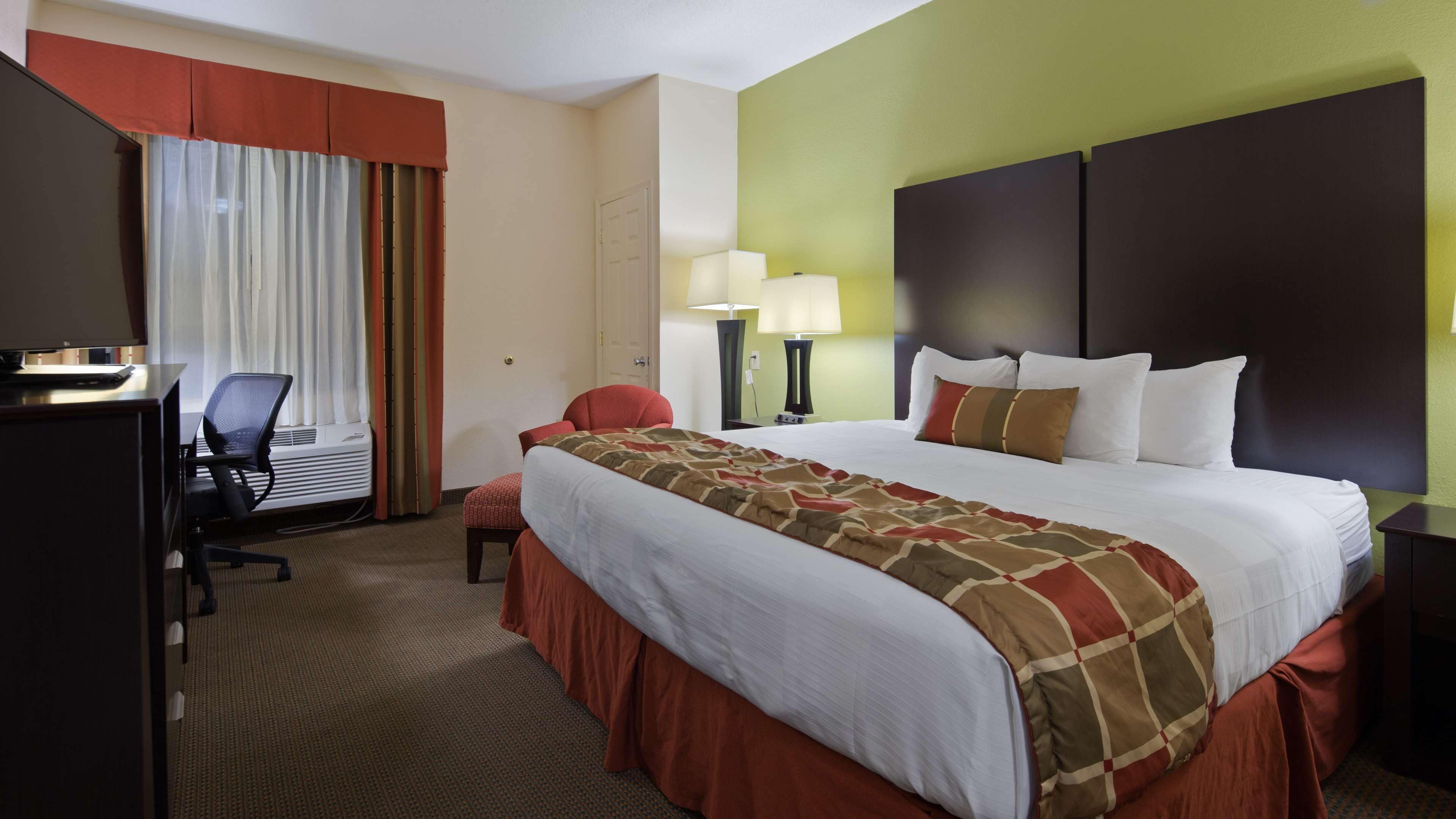 A hotel room with a large bed, a desk with a computer, a chair, and a window with curtains.
