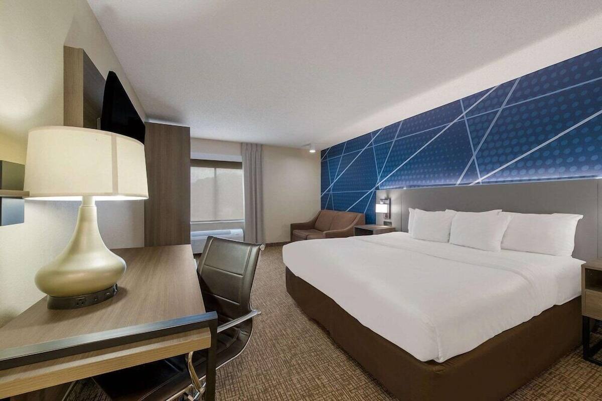 Una habitación de hotel con cama, escritorio, silla, lámpara y pared azul con diseño geométrico.