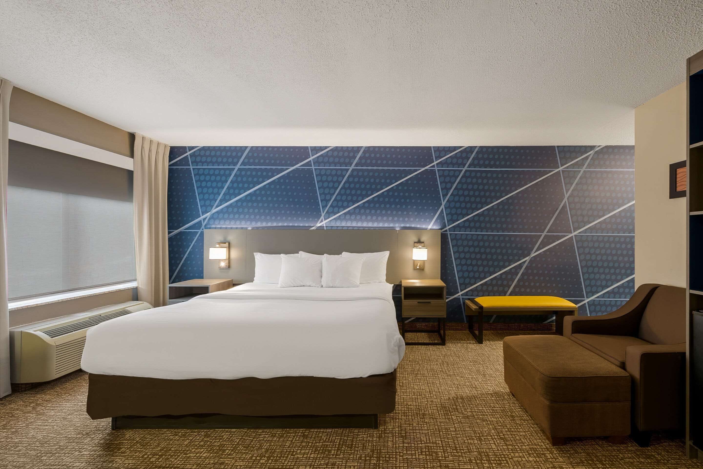 A hotel room with a large bed, two bedside tables with lamps, a chair, and a patterned wall.