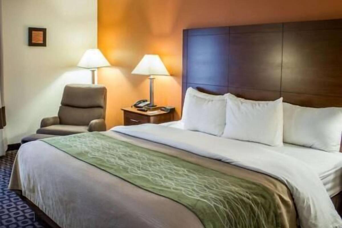 Una habitación de hotel con una cama, dos lámparas de noche, una silla, una mesita de noche y un cuadro en la pared.