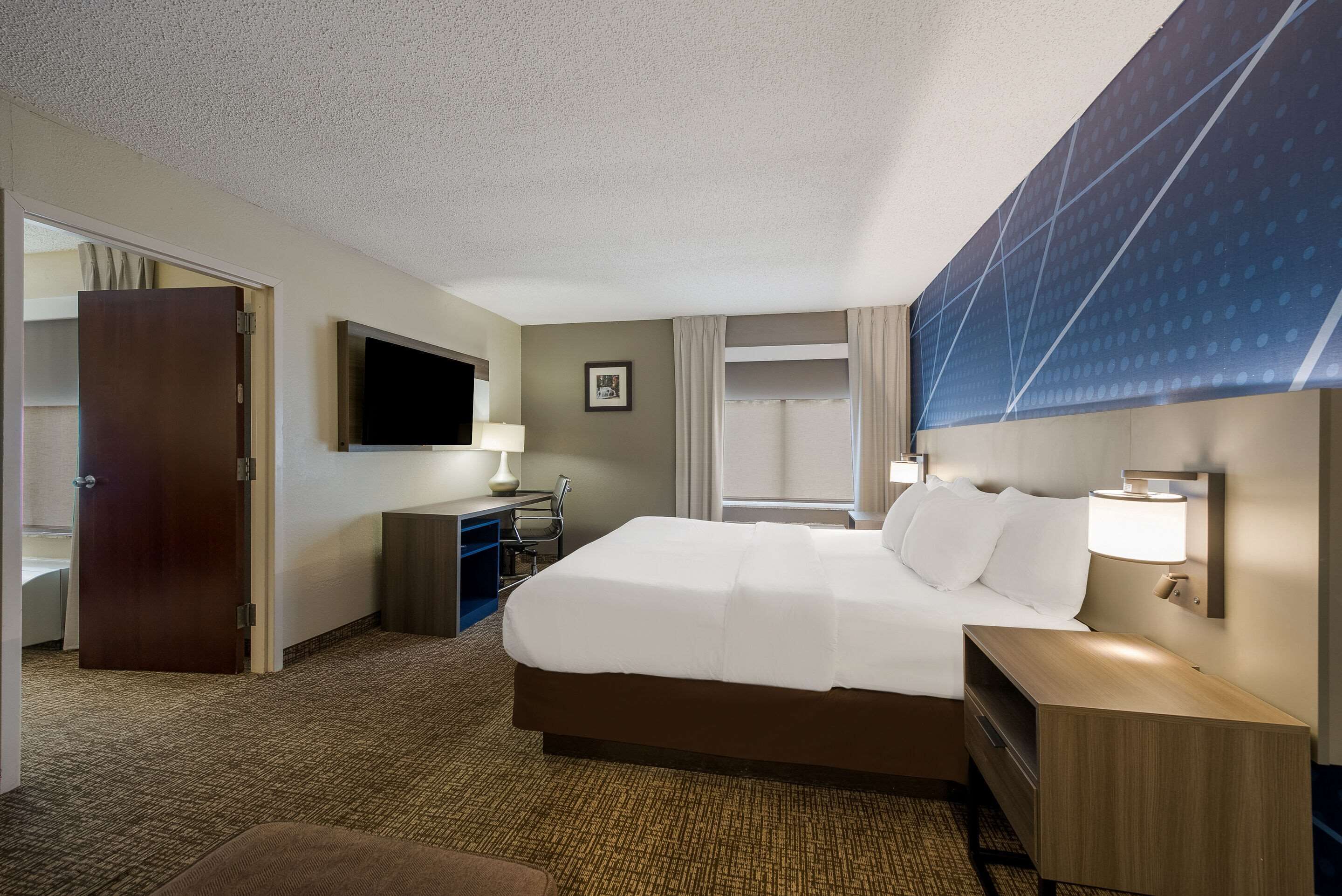 Habitación de hotel con una cama grande, un escritorio con lámpara, un televisor montado en la pared y una ventana con cortinas.