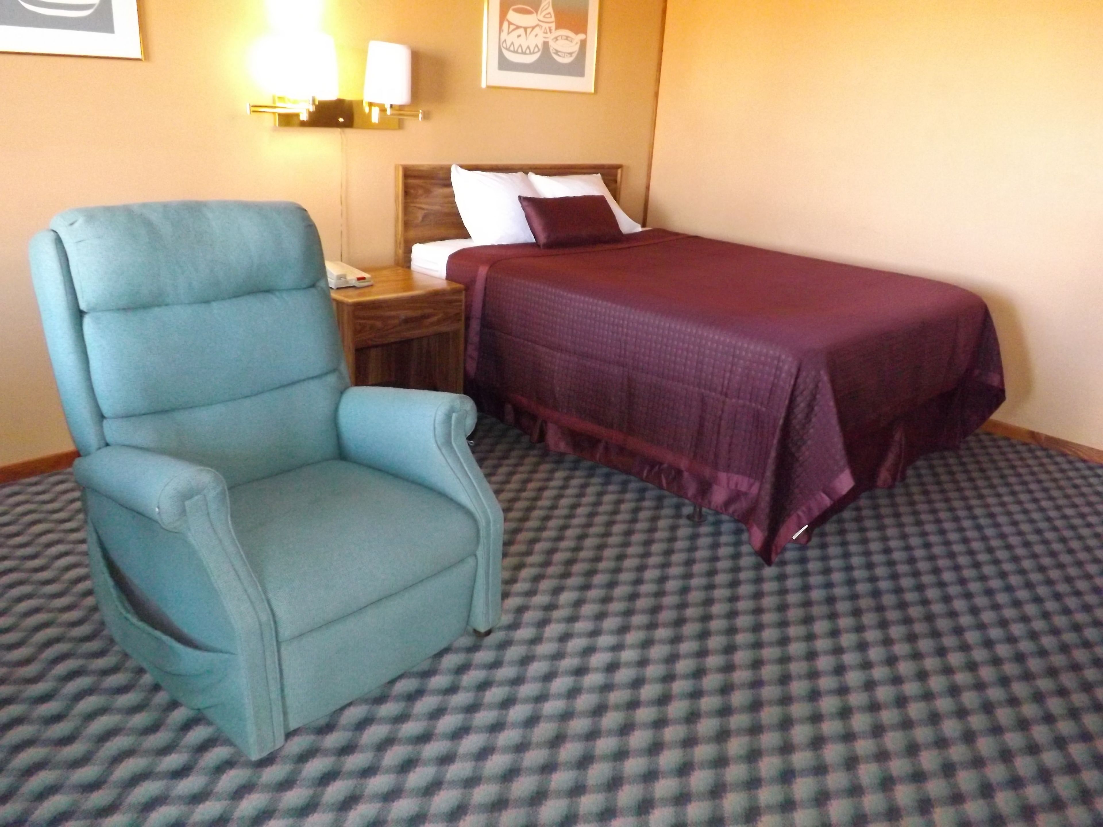 A hotel room with a teal armchair, a bed with a purple bedspread, a checkered carpet, and wall-mounted lamps.