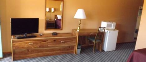 Habitación de hotel con tocador de madera, televisor, lámpara, silla y refrigerador.