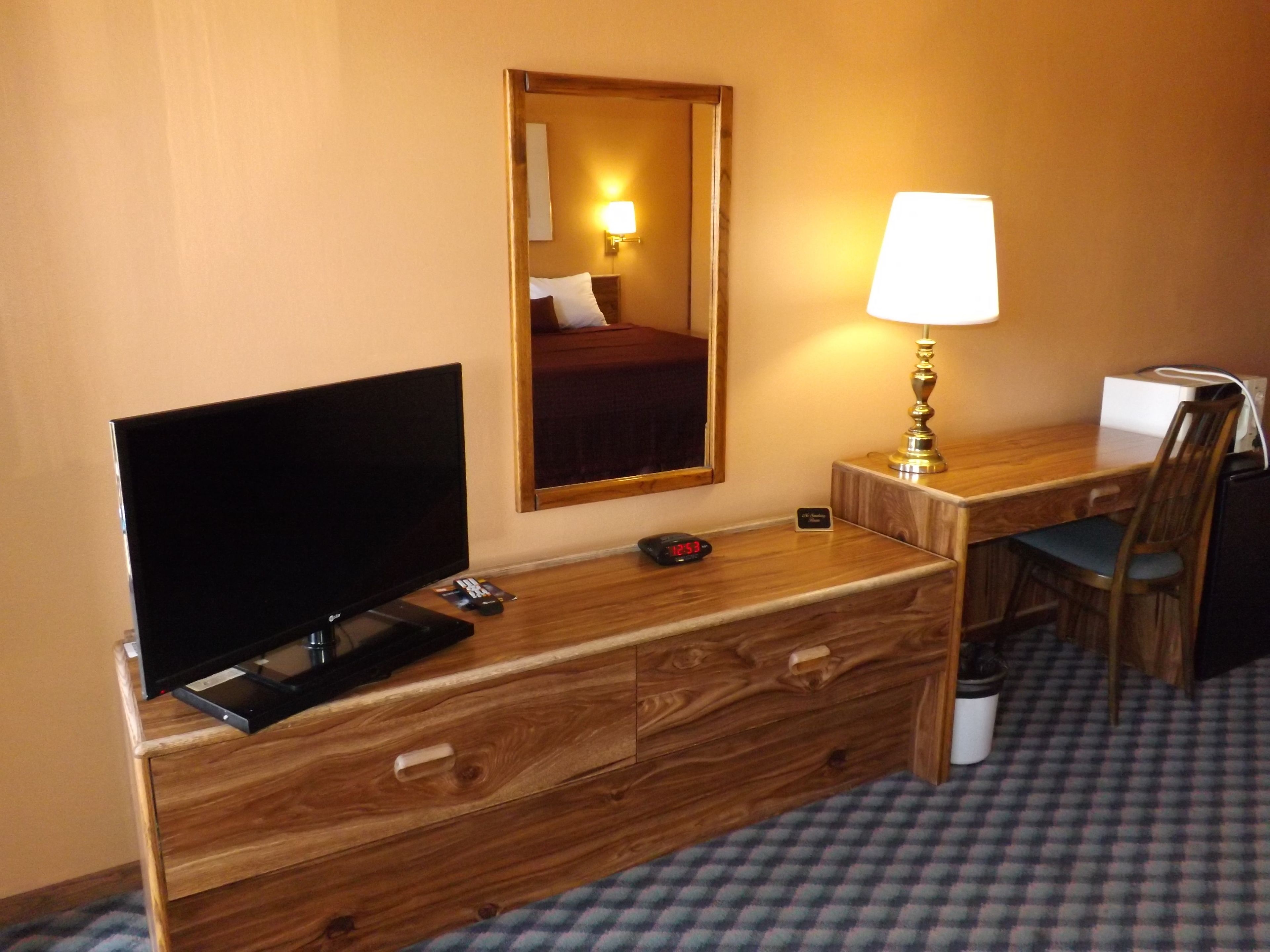A hotel room with a wooden desk, a television, a lamp, a chair, and a bed with a mirror above it.