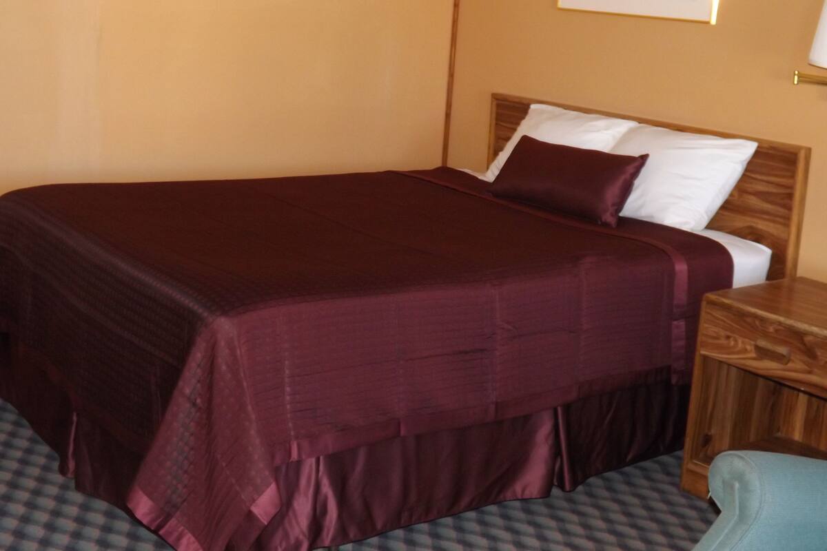 A single bed with a wooden headboard, a dark red bedspread, and a white pillow.