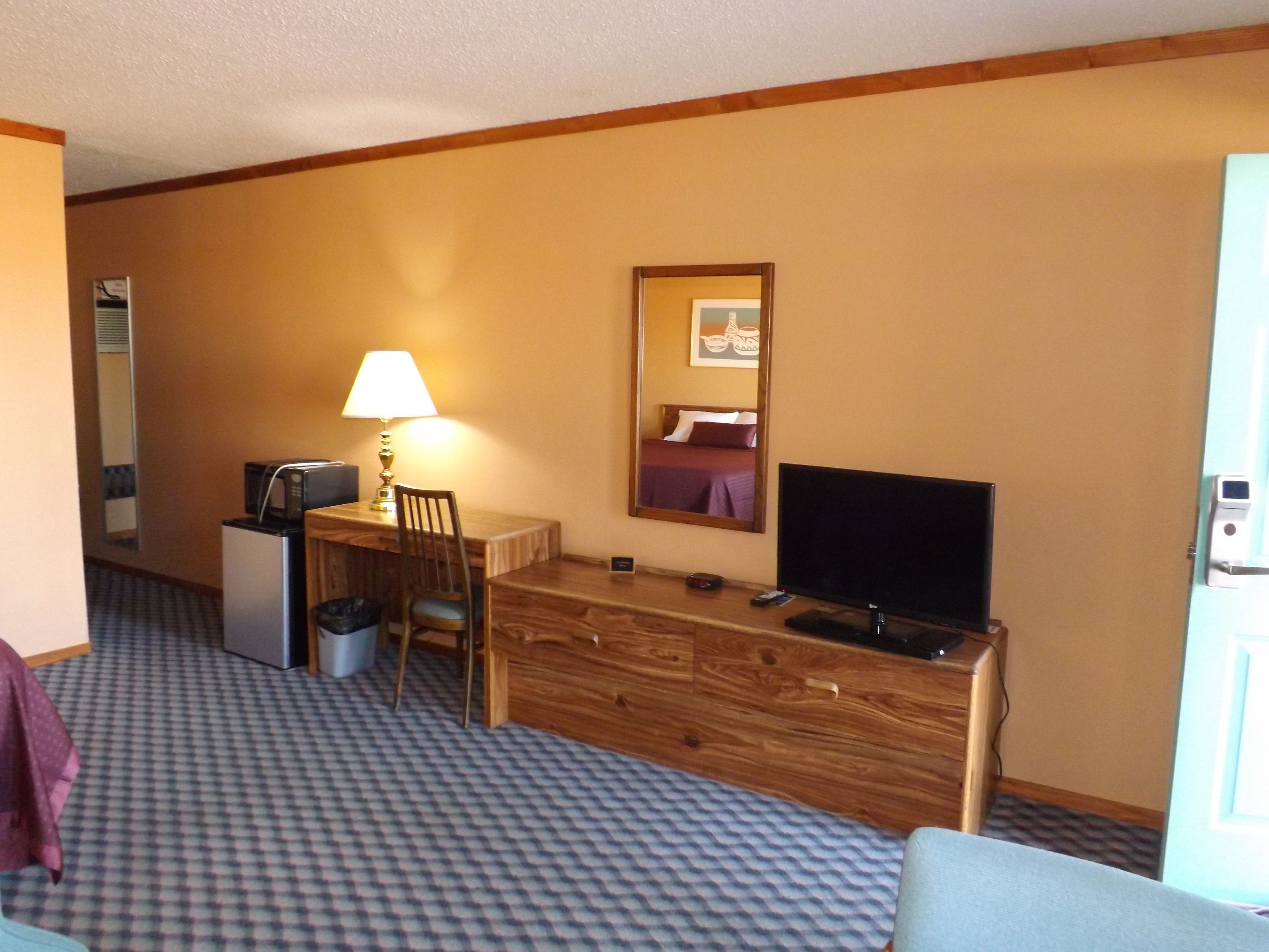 Quarto de hotel com uma escrivaninha de madeira, televisão, luminária, cadeira e cama.
