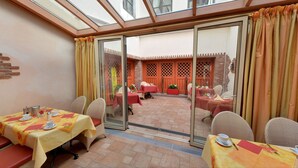 Un comedor con mesas dispuestas para dos, un ventanal de vidrio que da a un patio exterior y una pared de ladrillo con un diseño de celosía.
