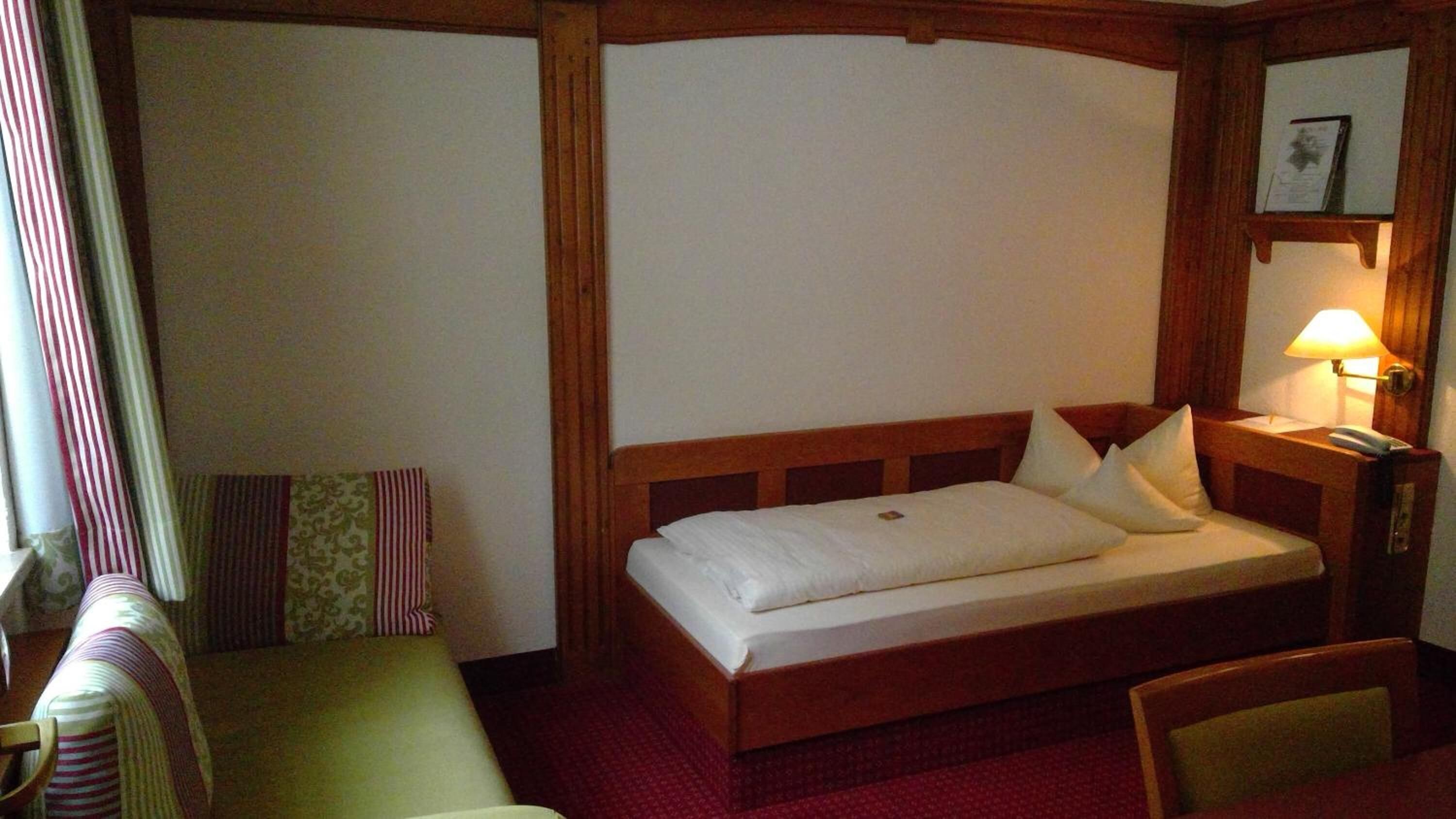 A hotel room with a bed, a bedside table with a lamp, a chair, and a window with curtains.